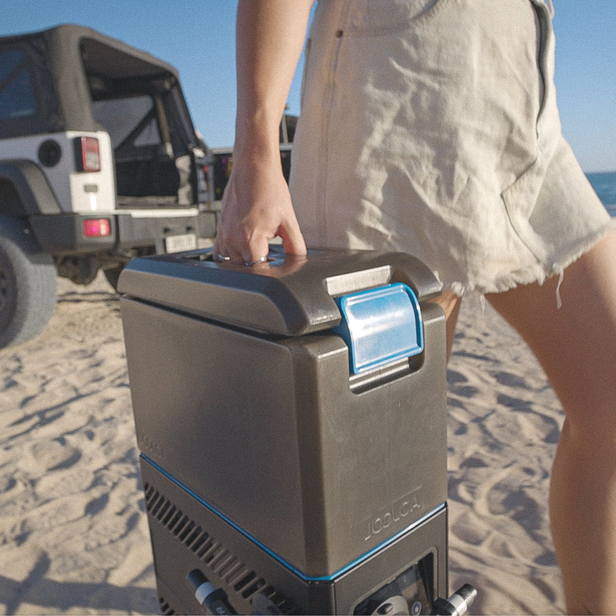 Person carrying a compact, portable hot water system on a sandy beach with a parked off-road vehicle in the background, emphasizing outdoor and off-grid use.