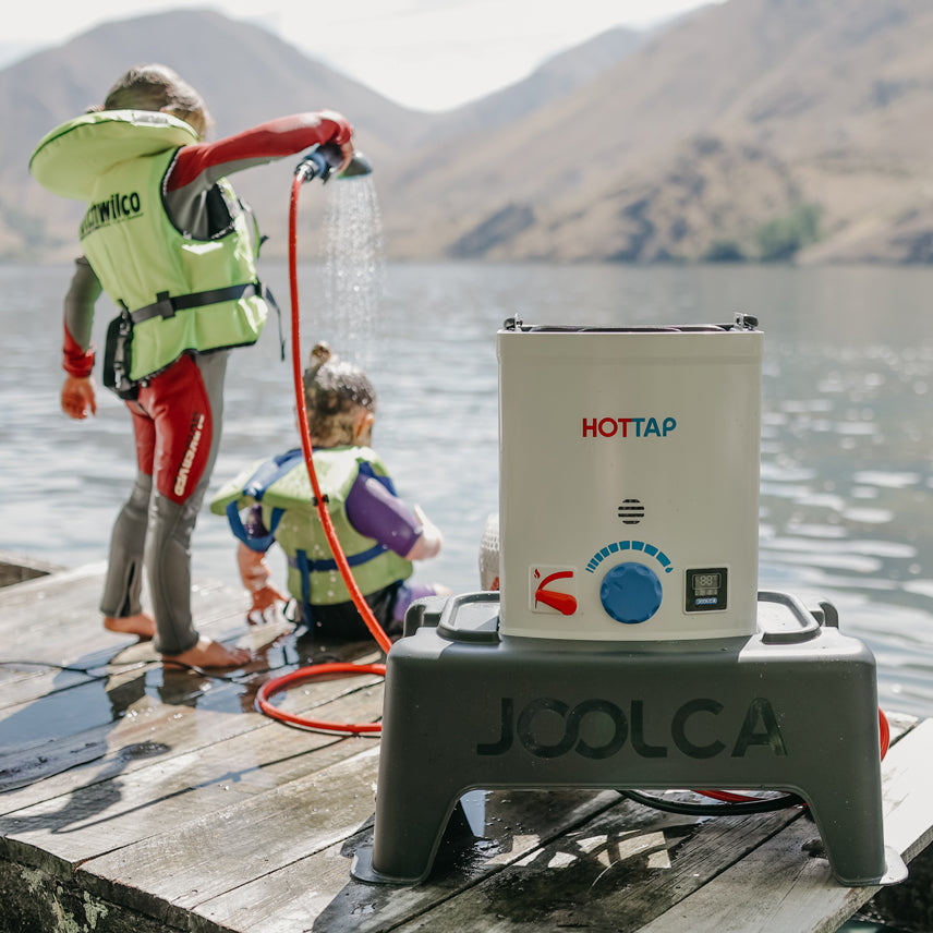 Portable outdoor hot water system setup on a dock beside a lake with two children in life jackets playing near the water, surrounded by natural mountainous scenery.