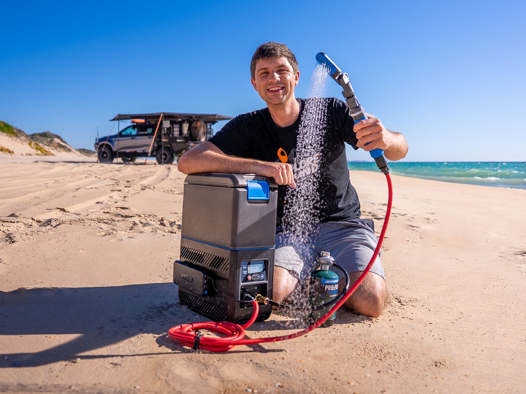 Person using a portable shower on a beach with a cooler and vehicle in the background