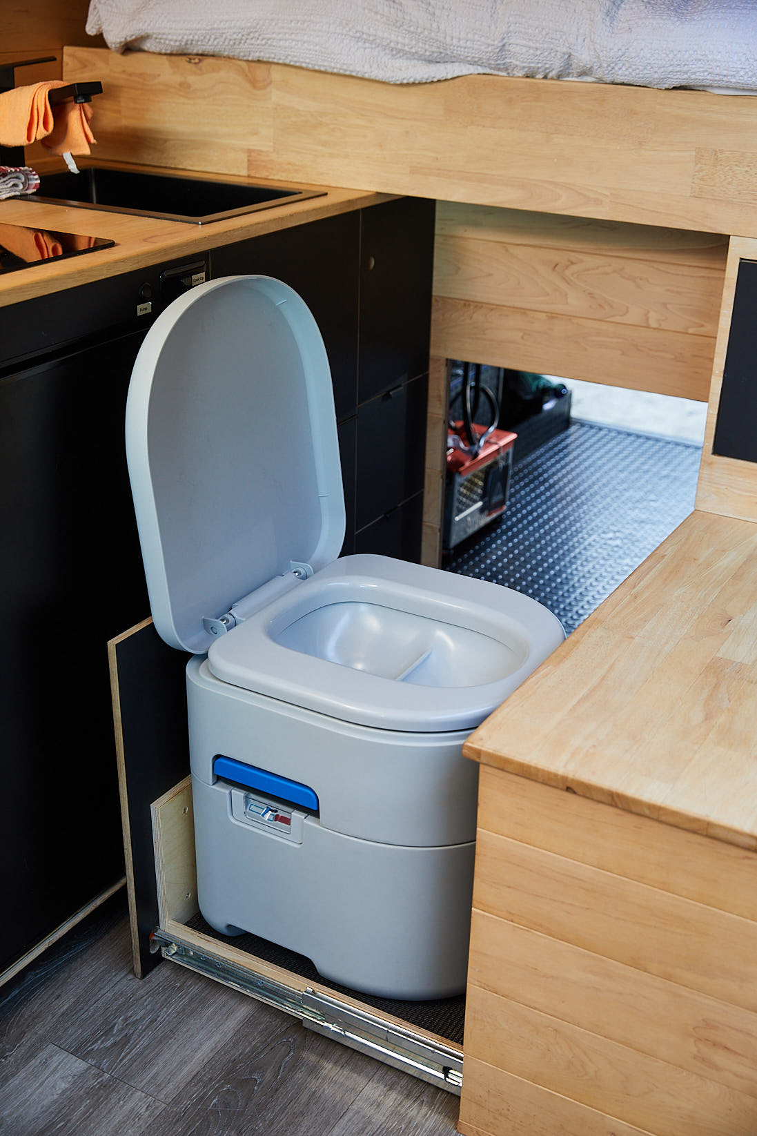 Portable gray composting toilet with open lid installed on a sliding track inside a wooden camper van interior near a black kitchenette and wooden countertops.