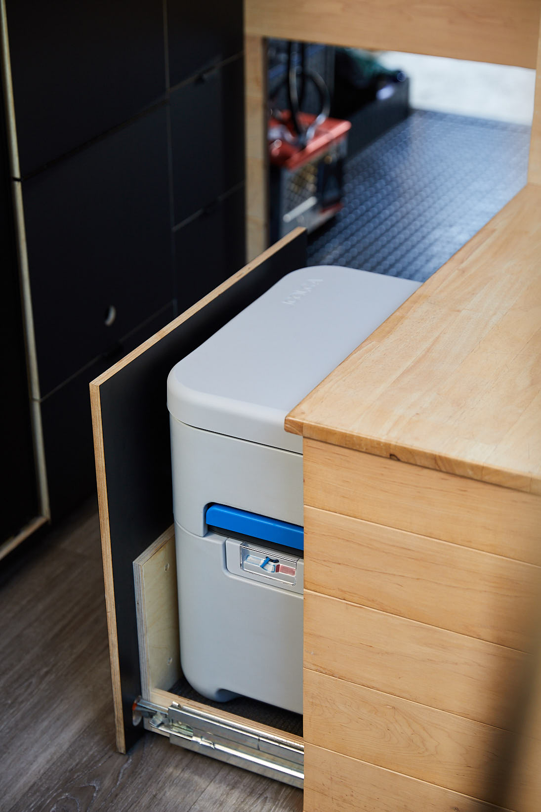 A compact portable toilet with a blue handle and control panel, installed on a sliding wooden rack between cabinetry and a wooden countertop.