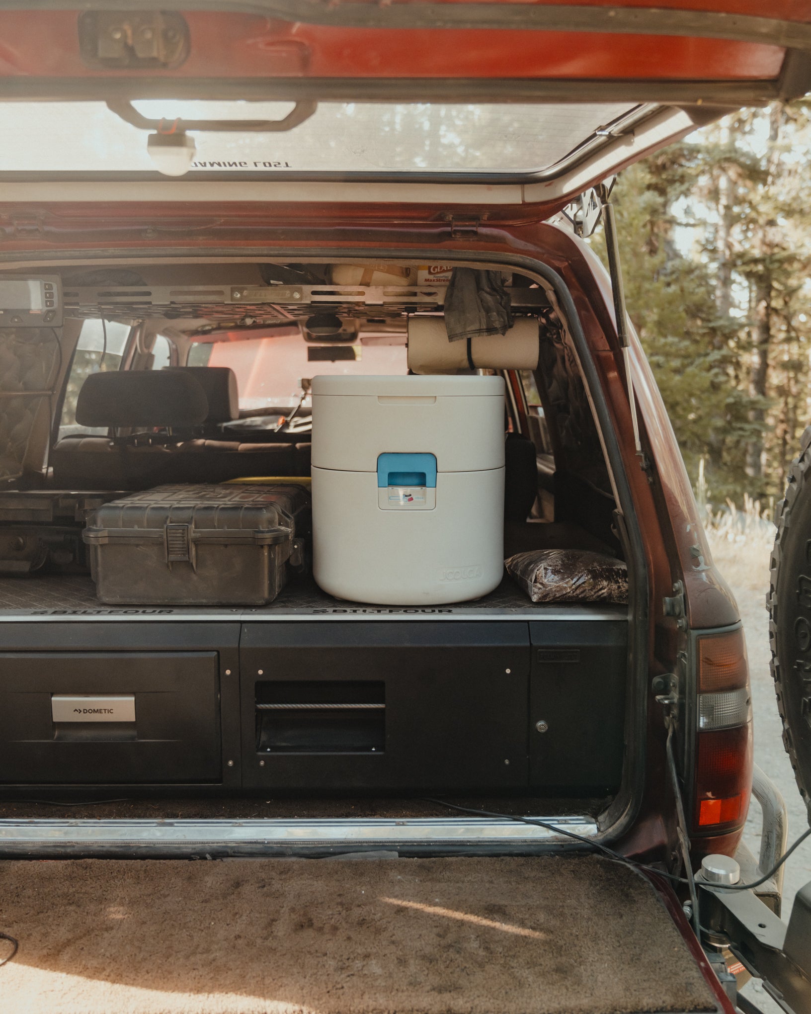Rear cargo area of a vehicle set up for outdoor adventure with a portable composting toilet, storage cases, and gear stored inside an open tailgate with a forest background.