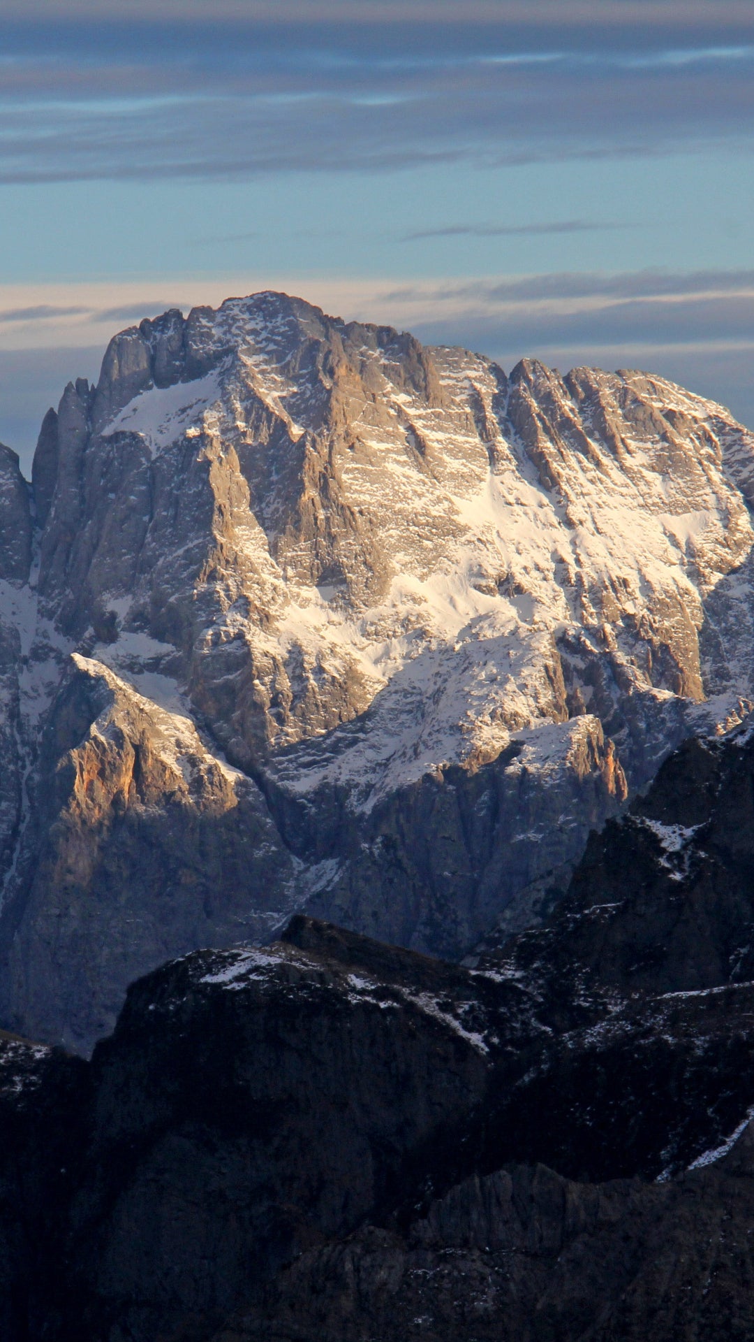 Snow-dusted mountain peaks with sharp rocky cliffs, lit by low-angle sunlight, under a pale blue sky with light clouds.