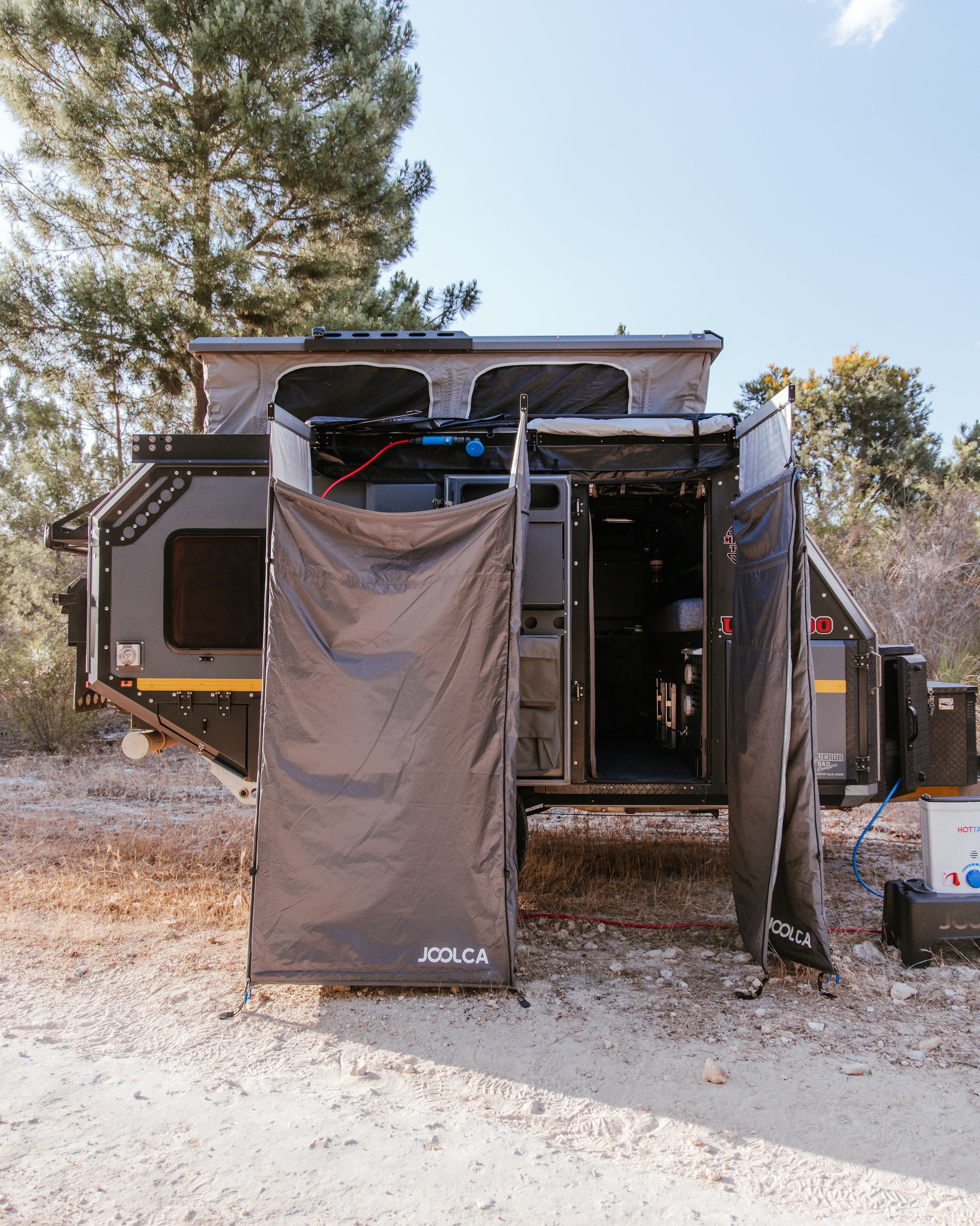 Two portable privacy tents set up outside a rugged off-grid camper trailer in a dry, wooded outdoor setting under a clear sky.