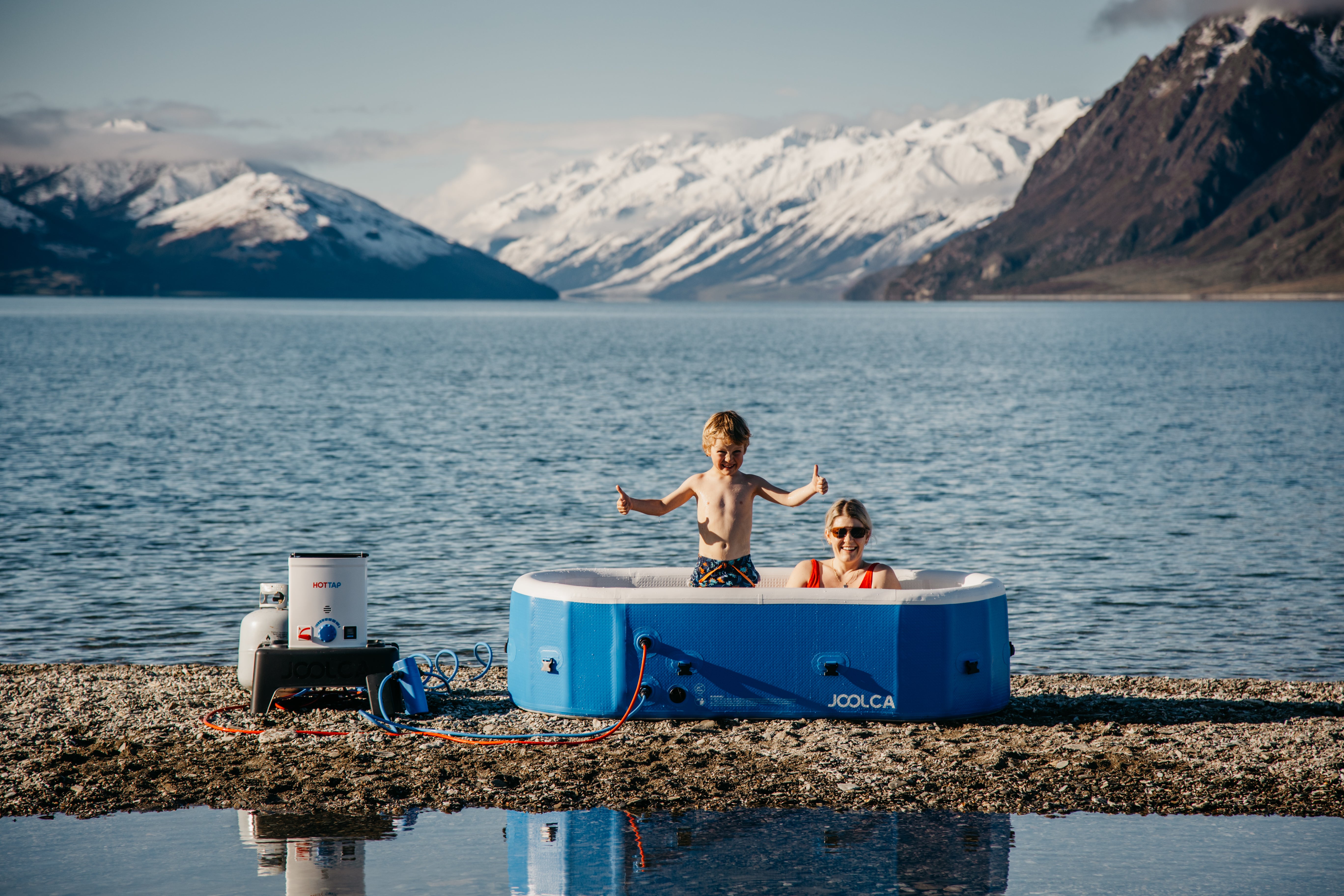 Two people in a blue portable hot tub by a lake with mountains in the background