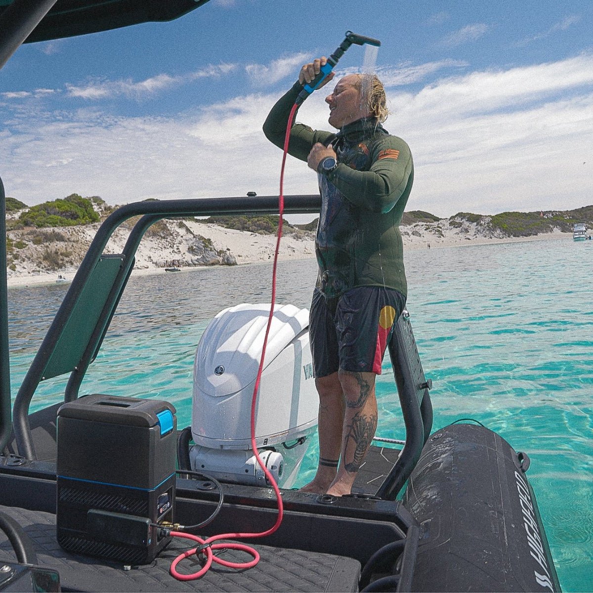 Man using Joolca HOTTAP Go portable hot water system on a boat for an outdoor shower, demonstrating off-grid hot water camping convenience by the clear turquoise water.