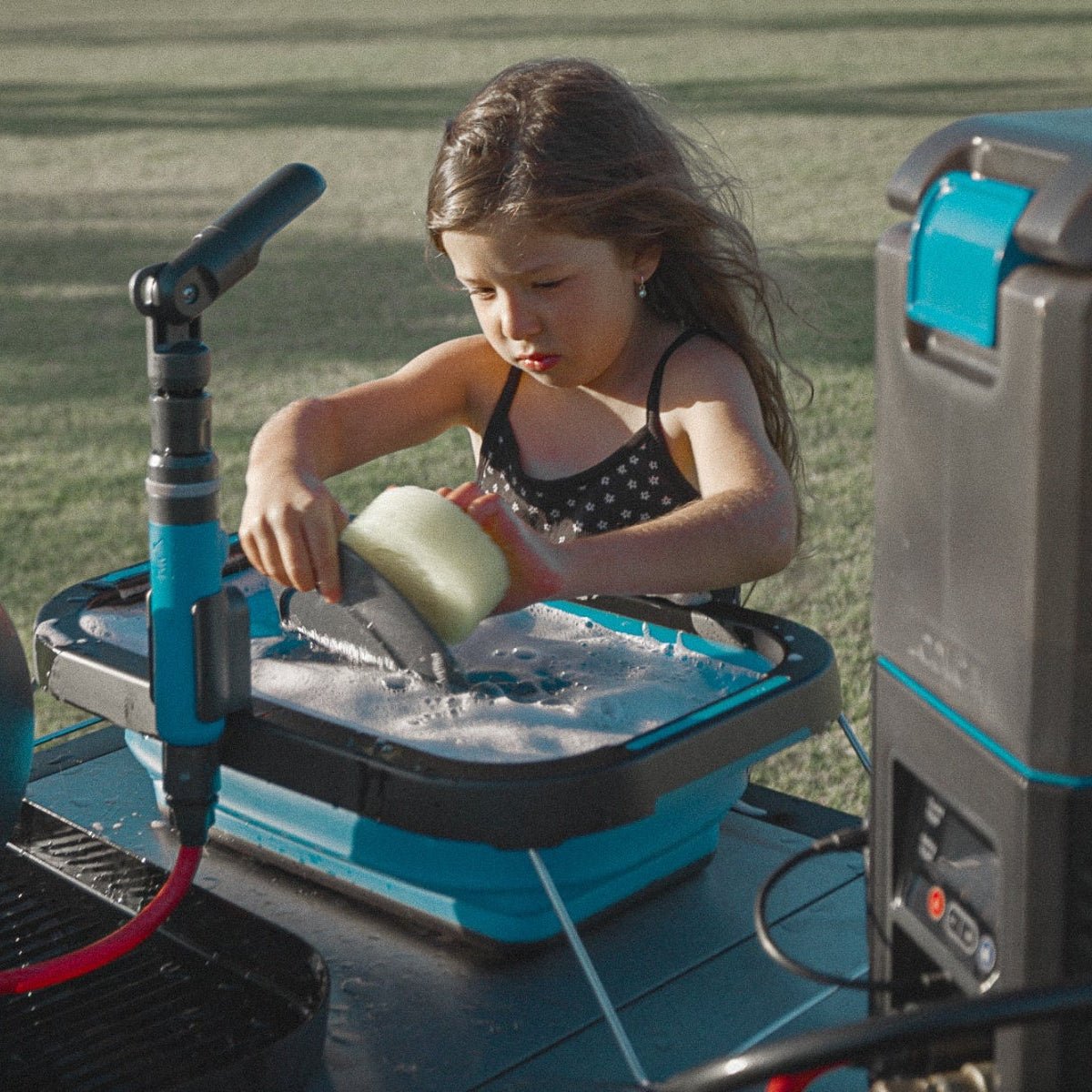 A young girl washing dishes outdoors using the HOTTAP Go portable instant hot water system with a blue and black collapsible basin and camping water heater by Joolca.