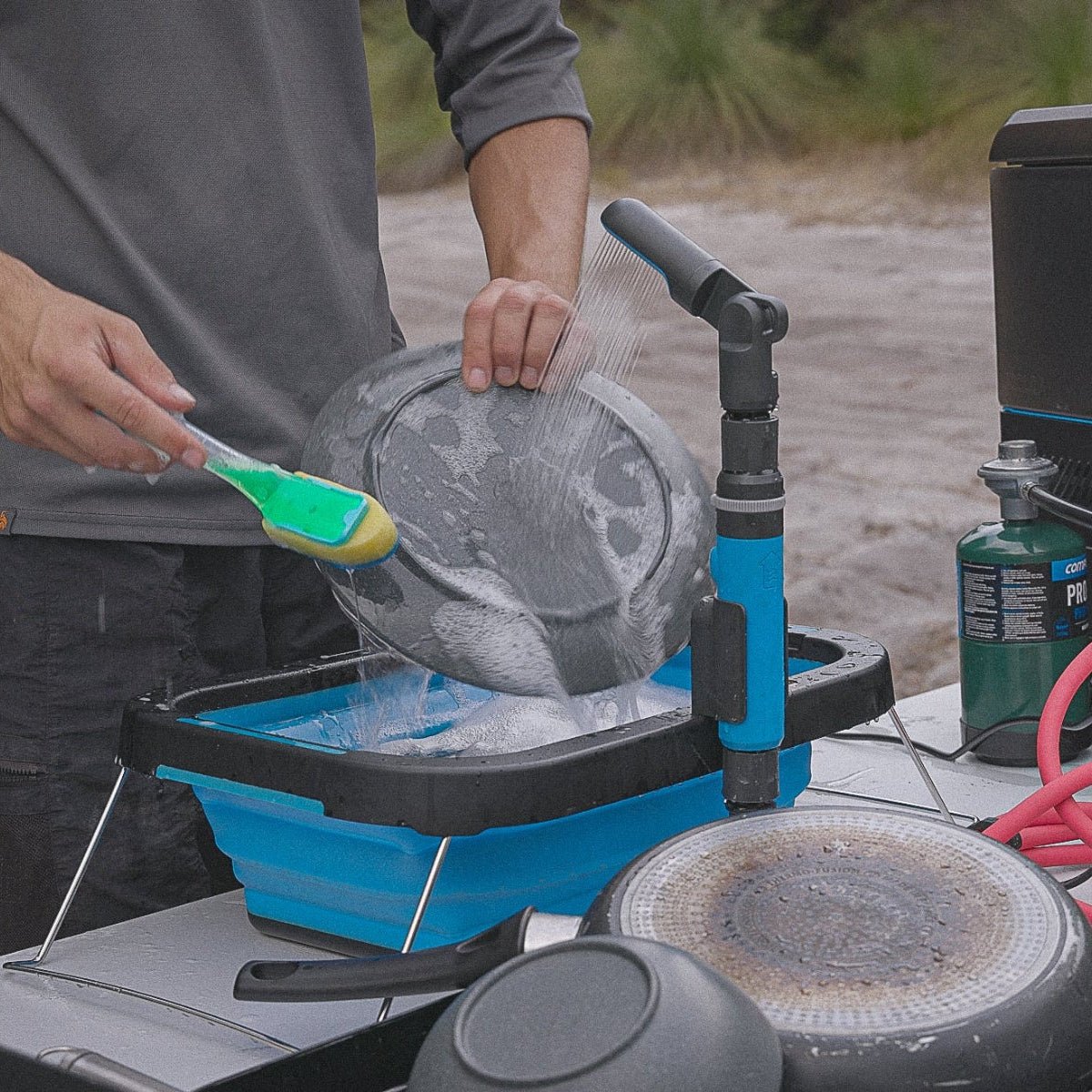 Outdoor scene showing a person washing a pan using a blue collapsible basin filled with water, with the Joolca HOTTAP Go portable instant hot water system providing a directed spray for easy cleaning during off-grid adventures.