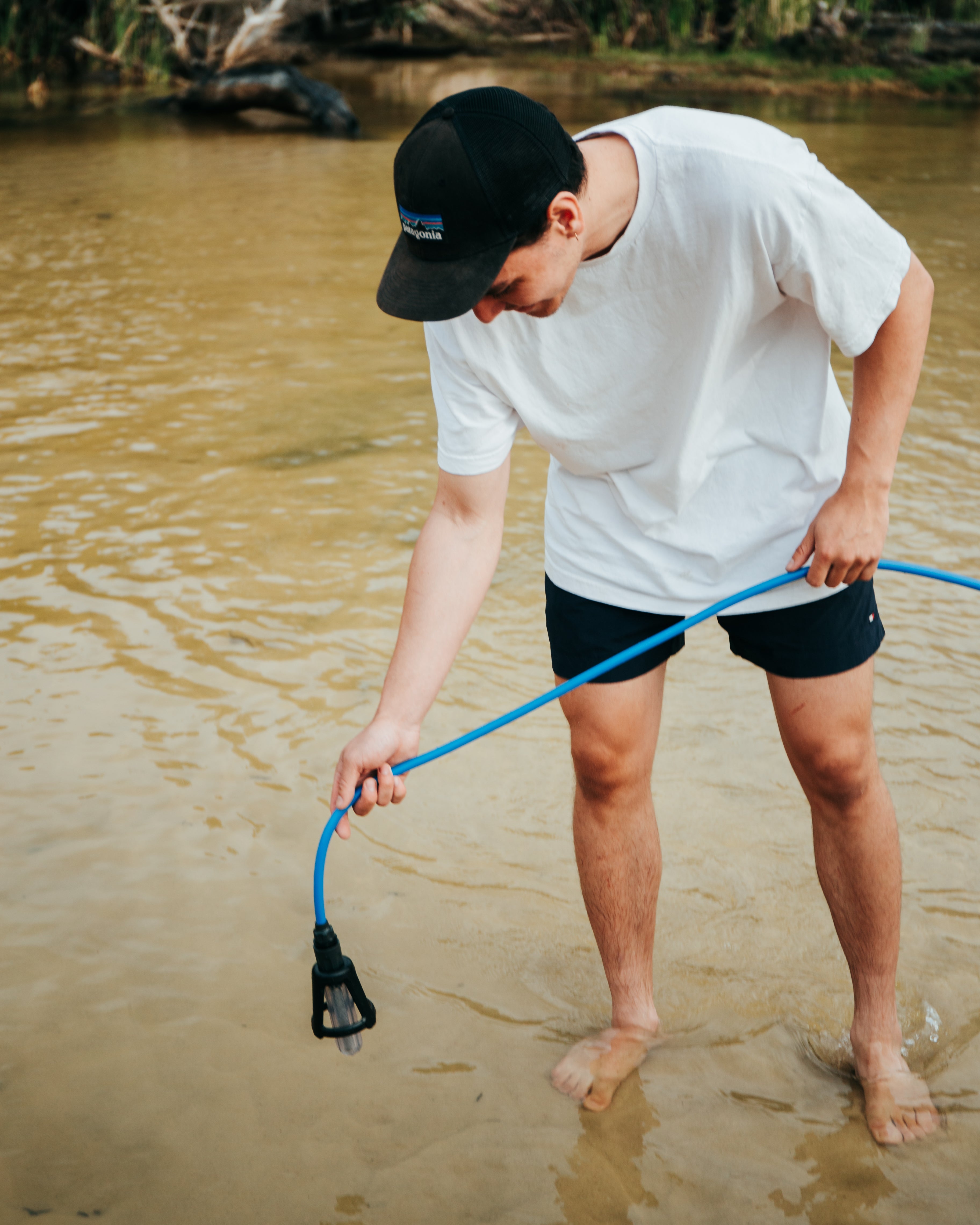 Person standing barefoot in shallow river water, holding a blue hose with a black nozzle, surrounded by natural greenery in the background.